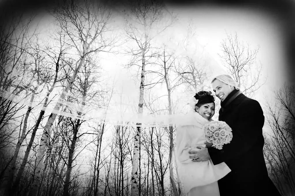 A joyful couple stands in a snowy birch forest, captured in an elegant black-and-white photograph. The bride holds a beautiful bouquet of roses, while her veil delicately floats in the winter breeze. Both are warmly dressed, with the groom in a dark coat. This image showcases the romantic and serene atmosphere that Maryse Creation, a distinguished wedding planner in Canada, can create. Known for crafting unforgettable wedding experiences, Maryse Creation falls under the wedding planners category on Mariage Elegant. The happy couple's expressions reflect the joy and harmony of their special day.