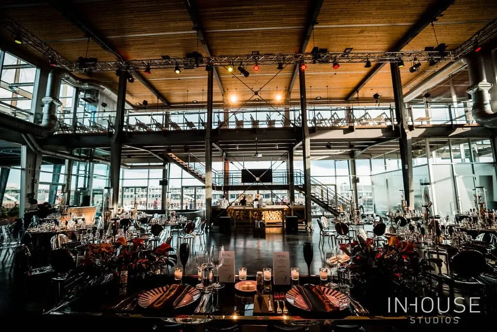 The image depicts a beautifully decorated event space suitable for weddings by Maryse Creation, a wedding planner categorized under Wedding Planners at Mariage Elegant, Canada. The venue features a spacious, modern interior with high ceilings and expansive windows that allow natural light to flood the room. Stylishly arranged round tables are set with elegant tableware, floral centerpieces, and candles, creating a romantic and sophisticated atmosphere ideal for a wedding reception. A prominent staircase in the background adds architectural interest, contributing to the venue's chic and contemporary aesthetic.