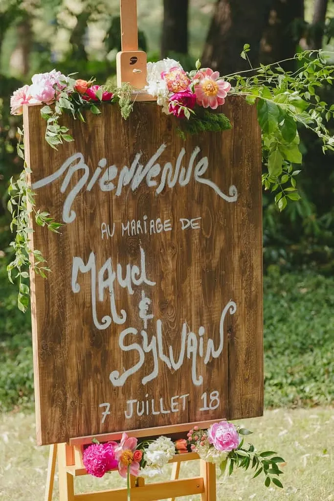 The image features a beautifully crafted wooden wedding sign adorned with vibrant pink and orange flowers, as well as lush greenery along the edges. The text on the sign is elegantly written in white and reads, 'Bienvenue au mariage de Maud & Sylvain, 7 Juillet 18.' This rustic and charming touch is likely part of the decor managed by Maryse Creation, a vendor in the Wedding Planners category, showcasing their skills in creating personalized and picturesque wedding elements for Mariage Elegant in Canada. The sunlit backdrop of trees adds a natural and serene ambiance, enhancing the welcoming atmosphere.