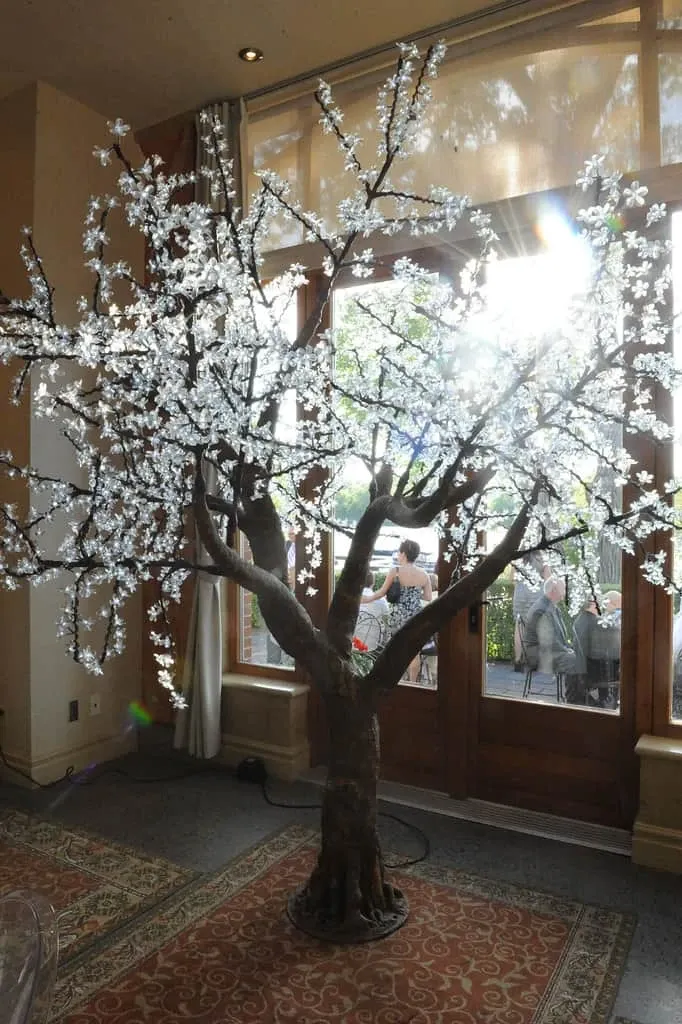 The image features an elegant decorative tree adorned with delicate, white blossoms, standing in a warmly lit room. Sunlight filters through large windows, casting a gentle glow on the tree, enhancing its ethereal beauty. This scene is set in a sophisticated indoor venue, showcasing an atmosphere suitable for weddings. The tree serves as a stunning centerpiece, possibly used in wedding decor to create a romantic and serene ambiance. The image relates to Maryse Creation, a vendor in the Wedding Planners category on Mariage Elegant, Canada, highlighting their capability to design exquisite and memorable wedding settings.