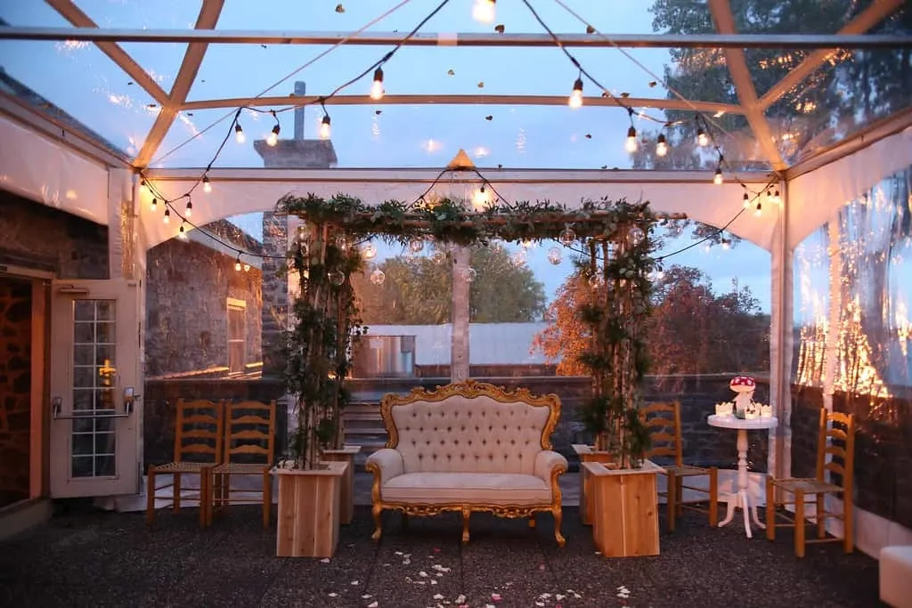 The image showcases an elegantly decorated wedding venue by Maryse Creation, a wedding planner under the Wedding Planners category at Mariage Elegant, Canada. The scene features a luxurious cream-colored loveseat with ornate gold detailing, positioned under a transparent tent structure. String lights are gracefully draped across the tent, casting a warm and inviting glow. Surrounding the loveseat are lush green garlands with hanging floral arrangements, adding a touch of natural beauty. Wooden chairs are placed to the sides, and a small white table holds a beautifully decorated cake, enhancing the romantic ambiance of this wedding setting.