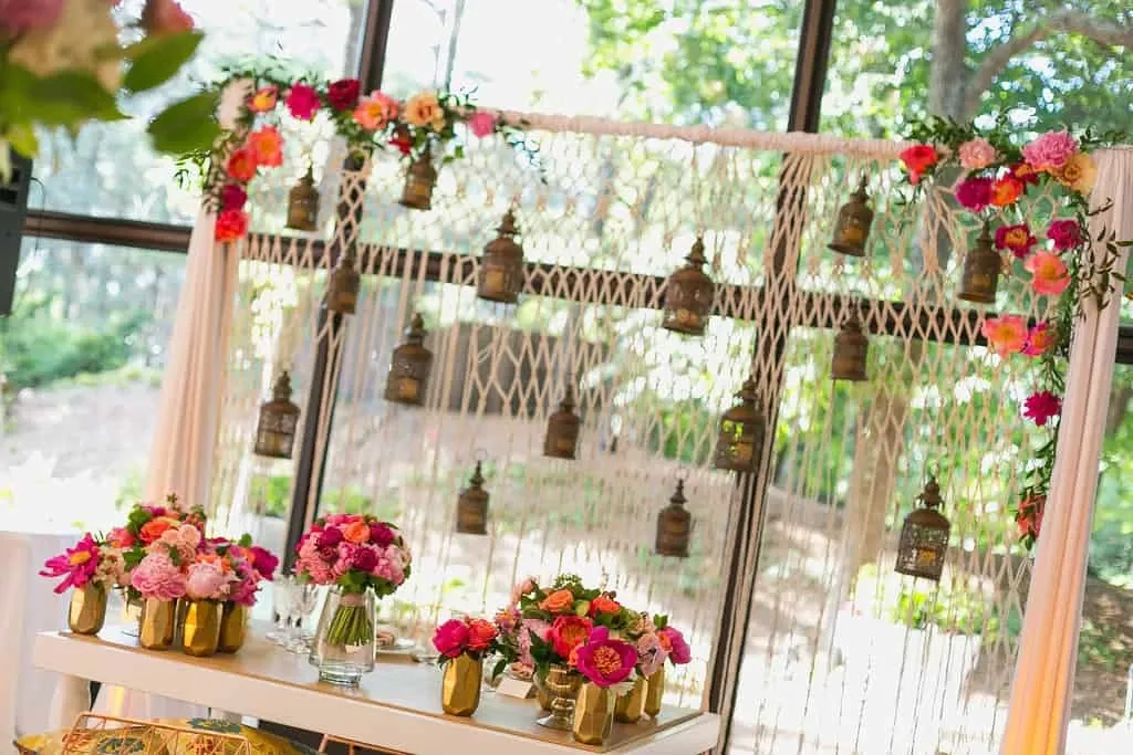 The image shows a beautifully decorated wedding backdrop created by Maryse Creation, categorized under wedding planners on Mariage Elegant, Canada. This arrangement features an intricate macramé backdrop adorned with hanging lanterns, adding a vintage touch. Vibrant floral arrangements with red, pink, and orange flowers sit elegantly on a shelf below, placed in golden vases. The scene exudes a romantic and sophisticated ambiance, ideal for a memorable wedding setting. Sunlight filters through large windows, highlighting the natural beauty and serene outdoor view.