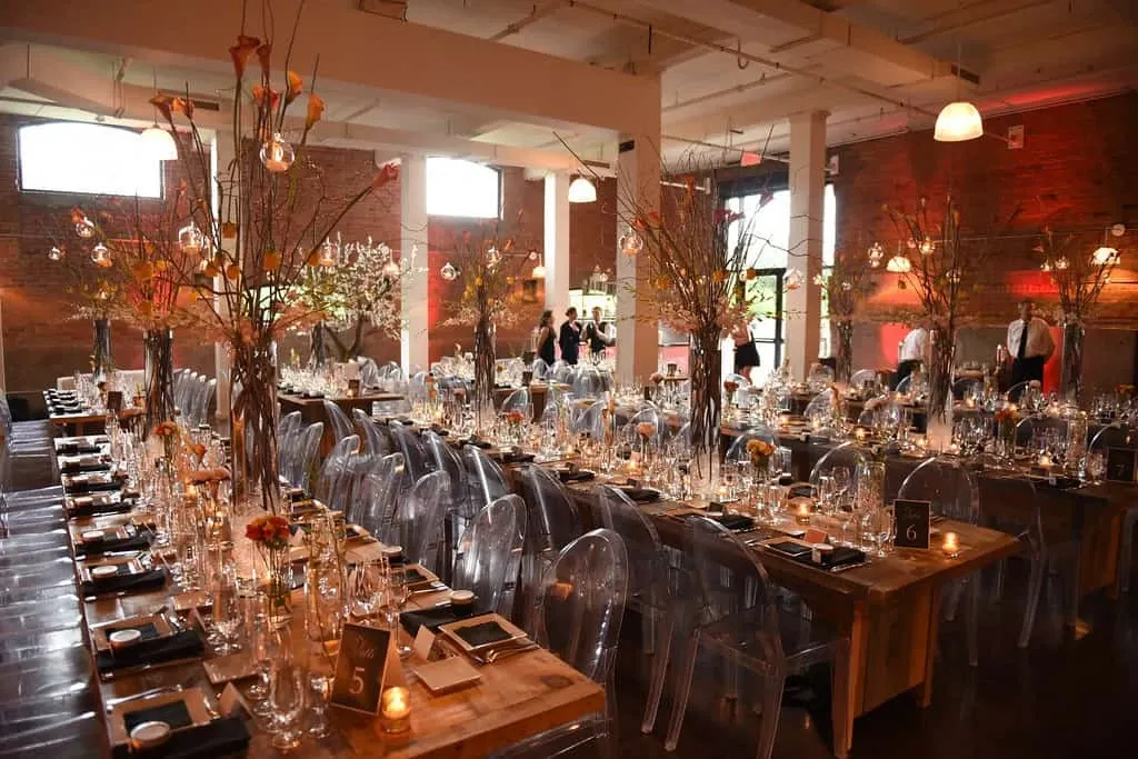 The image showcases a beautifully arranged wedding reception venue by Maryse Creation, a wedding planner listed under the Wedding Planners category on Mariage Elegant, Canada. The setting features long wooden tables elegantly decorated with tall vases filled with branches and delicate flowers, creating a sophisticated and natural ambiance. Each table is set with transparent chairs, gleaming glassware, and neatly arranged place settings, including black napkins and menu cards. Soft candlelight adds a warm glow to the scene, enhancing the romantic atmosphere. The industrial-style venue has exposed brick walls and ample lighting, contributing to an inviting and chic environment for a wedding celebration.