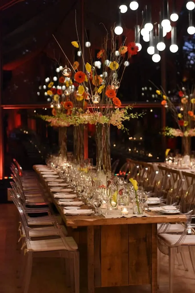 The image shows a beautifully decorated wedding reception table designed by Maryse Creation, a wedding planner listed in the Mariage Elegant vendor directory in Canada. The long wooden table is adorned with tall floral centerpieces featuring vibrant orange and yellow flowers, interspersed with delicate hanging glass candle holders emitting a warm glow. Crystal-clear wine glasses, pristine white plates, and neatly folded napkins are meticulously arranged along the table. Elegant transparent chairs surround the setup, and overhead, modern spherical lights add to the sophisticated ambiance, reflecting the expertise of Maryse Creation in creating enchanting wedding atmospheres.