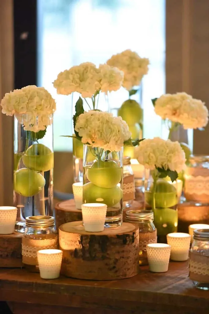 This elegant image showcases a beautifully arranged wedding centerpiece by Maryse Creation, categorized under Wedding Planners at Mariage Elegant, Canada. The display features tall glass vases filled with crisp green apples and topped with white hydrangeas, creating a fresh, sophisticated look. The vases are artfully placed on rustic wooden slices, surrounded by softly glowing candles in delicate holders. This arrangement exemplifies a blend of natural elements and romantic ambiance, ideal for a stylish and memorable wedding setting.