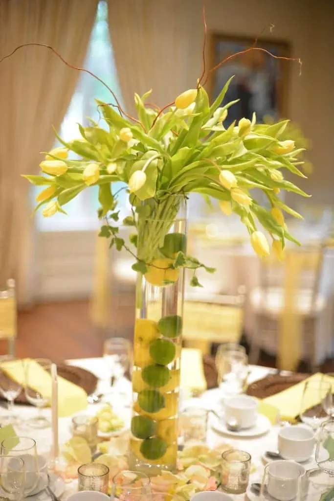 This image showcases a beautifully arranged wedding reception table planned by Maryse Creation, categorized under Wedding Planners. At the center is a tall, elegant glass vase filled with layered lemons and limes, topped with a stunning bouquet of yellow tulips and green foliage. The table is set with fine china, glassware, and neatly folded napkins, surrounded by gold Chiavari chairs, creating an inviting and sophisticated atmosphere. The soft draping curtains in the background add a touch of elegance, perfectly illustrating the meticulous detail and artistry in Maryse Creation's wedding planning.
