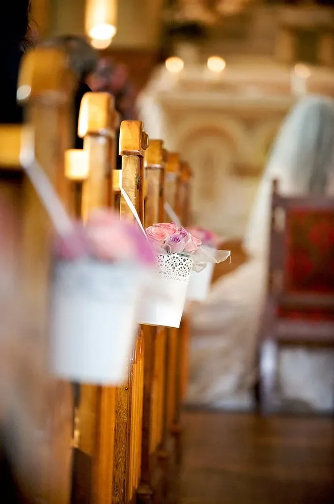 The image shows a beautifully decorated church aisle with wooden pews adorned with small white buckets filled with pink roses, creating an elegant and romantic atmosphere. In the blurred background, a bride in a flowing white gown is visible, enhancing the wedding setting. This image represents the high-quality services provided by Maryse Creation, a top wedding planner listed under the Wedding Planners category on Mariage Elegant, Canada. The attention to detail in floral arrangements highlights the expertise of Maryse Creation in creating memorable wedding moments.