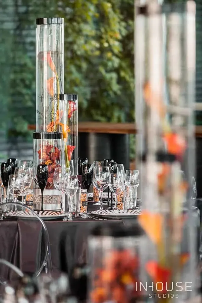 This image features an elegantly arranged wedding reception table designed by Maryse Creation, a renowned vendor in the Wedding Planners category at Mariage Elegant, Canada. The table is adorned with tall cylindrical vases filled with vibrant orange and red foliage, creating a striking centerpiece. Surrounding the vases are neatly arranged wine glasses and silverware, reflecting the natural light. The backdrop includes lush greenery, adding a fresh and sophisticated ambiance to the setting. The setup exemplifies a luxurious and stylish wedding theme, showcasing Maryse Creation's expertise in event planning.