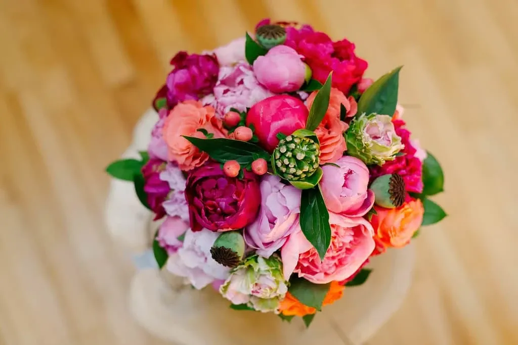 A vibrant bridal bouquet featuring an assortment of bright, colorful flowers including pink peonies, orange and red roses, and green foliage, artistically arranged on a light wooden surface. This beautiful floral arrangement is representative of Maryse Creation, a featured vendor in the Wedding Planners category on Mariage Elegant, Canada. The bouquet is ideal for a cheerful and vibrant wedding theme, showcasing the vendor's expertise in elegant and creative floral designs for weddings.