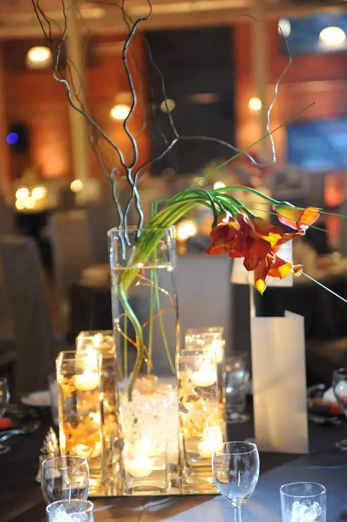 A beautifully arranged wedding table centerpiece by Maryse Creation, a wedding planner under the Wedding Planners category on Mariage Elegant, Canada. The arrangement features a tall, clear glass vase filled with elegant, twisting branches and vibrant orange calla lilies. Surrounding the central vase are smaller glass votives with flickering candles, casting a warm glow over the table. The scene is set in an ambient venue with soft lighting, creating a romantic and sophisticated atmosphere, ideal for a memorable wedding celebration.