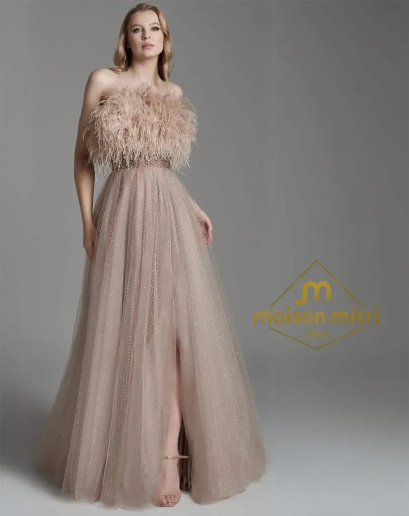 A woman poses in a chic, floor-length champagne gown featuring a strapless design with a feathered bodice and a flowing tulle skirt. This elegant dress, highlighted by subtle shimmering details, embodies sophistication and glamor. The background is a simple gray, allowing the dress's textures to stand out. The 'Maison Mitri' logo is prominently displayed, positioning this attire within their wedding tuxedo collection at Mariage Elegant in Canada.