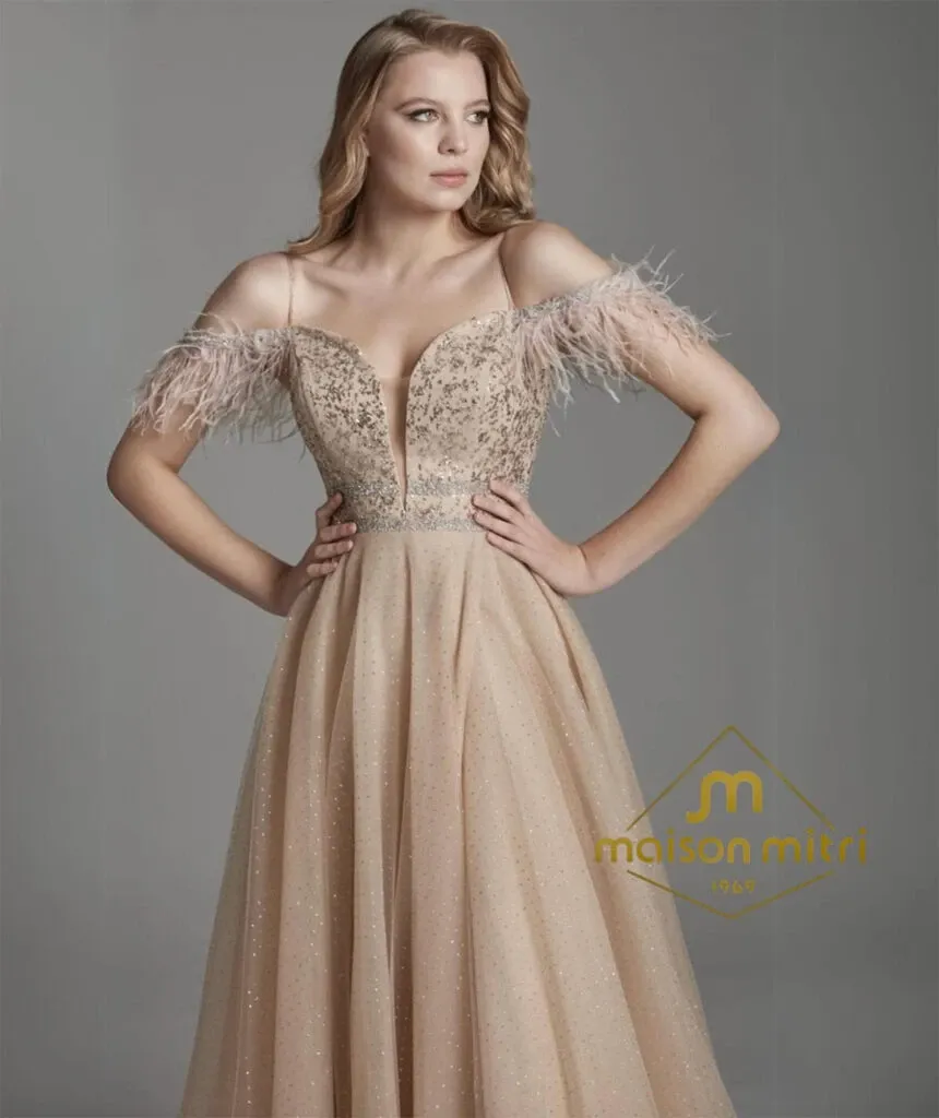 A woman models an elegant, off-the-shoulder gown adorned with intricate beadwork and delicate feather details at the sleeves, set against a simple gray backdrop. The gown features a deep plunging neckline and a full, flowing skirt, offering a blend of classic and contemporary bridal fashion. The image includes the logo and name of Maison Mitri in the bottom right corner, a vendor specializing in wedding tuxedos, indicating a high-quality, reputable brand. This image serves as a representation of the sophisticated wedding attire options available for the discerning couple at Mariage Elegant, Canada.