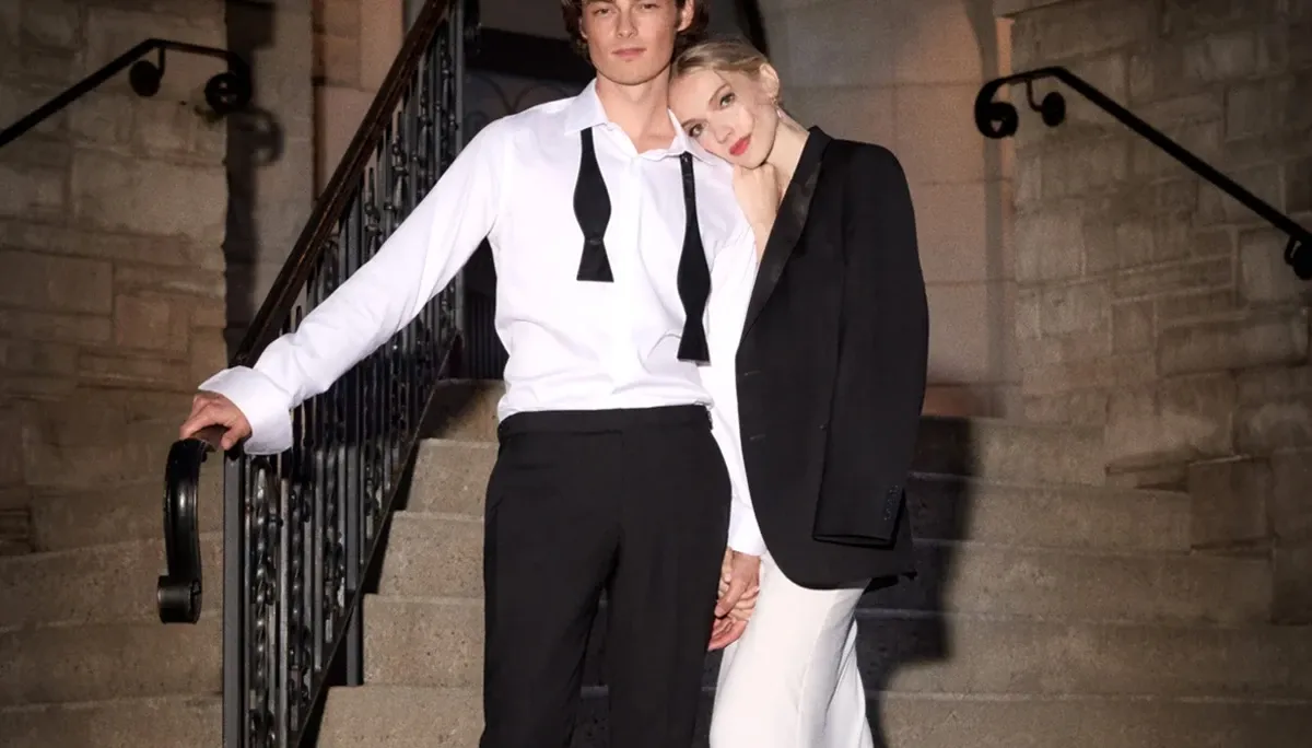 A couple stands on an elegant stone staircase, illuminated softly by ambient lighting, creating a romantic and sophisticated setting. The man, representing Tip Top in the Wedding Tuxedo category, is dressed in a stylish ensemble featuring black formal trousers, a crisp white shirt with the bowtie untied and hanging around his neck, creating a relaxed yet polished look. The woman beside him leans gently on his shoulder, wearing a chic black blazer over her white outfit, adding to the formal aesthetic. This image captures an intimate and fashionable moment, ideal for a wedding planned through Mariage Elegant in Canada.