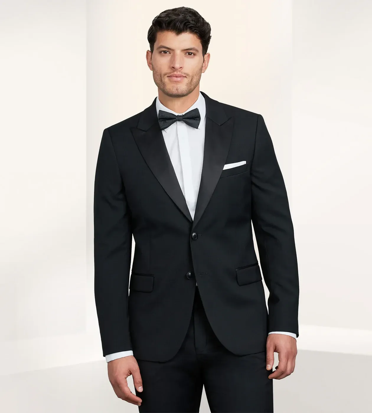 A groom wearing a sleek black tuxedo stands confidently in a minimalistic setting. The tuxedo, provided by the vendor Tip Top under the Wedding Tuxedo category, features a classic design with a satin lapel, paired with a crisp white dress shirt and a black bow tie. The setting suggests a formal and elegant atmosphere suitable for a wedding occasion.