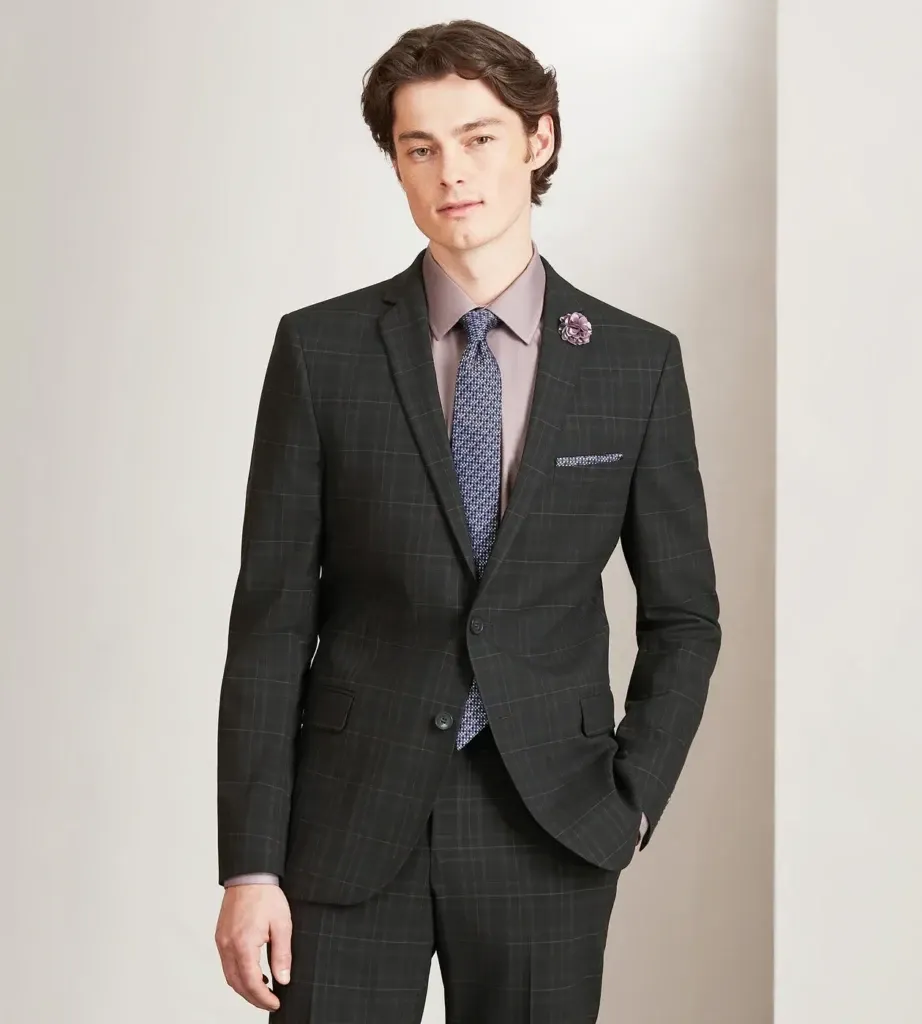 A well-dressed man is modeling a sleek, dark plaid tuxedo from the vendor Tip Top, part of their wedding tuxedo collection. The ensemble features a tailored jacket with a single-breast design and matching trousers. A light pink dress shirt is paired with a textured blue tie and a matching pocket square. A subtle floral boutonniere adds a touch of elegance, perfect for a stylish wedding look. The setting is minimalistic with a soft, neutral-colored background, highlighting the sophistication of the attire. This tuxedo option is ideal for those seeking a modern yet classic look for their wedding day.