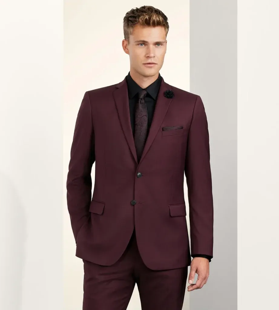 A groom is wearing a sleek, burgundy wedding tuxedo from the vendor Tip Top, categorized under Wedding Tuxedo. The tuxedo features a tailored fit with a single-breasted jacket and notched lapels, complemented by two black buttons. The ensemble is paired with a black dress shirt and a matching burgundy tie, adding an elegant contrast to the overall look. A dark pocket square is neatly tucked into the chest pocket, enhancing the sophisticated style ideal for a formal wedding setting. The backdrop consists of a minimalist, light-colored wall that accentuates the rich hue of the tuxedo.