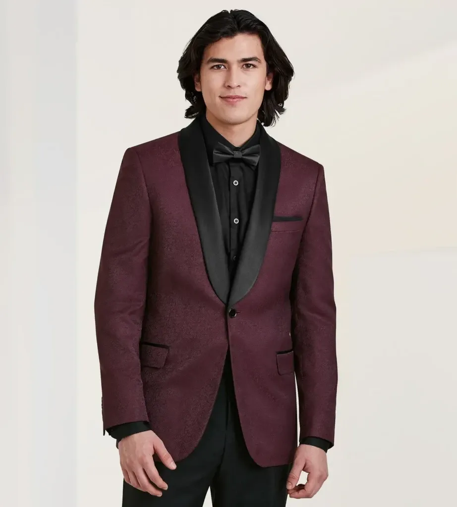 A young man is modeling a sophisticated wedding tuxedo from the vendor Tip Top, featured in the Wedding Tuxedo category. The tuxedo jacket is a rich burgundy color with a subtle textured pattern, complemented by a sleek black satin shawl lapel. He is wearing a black dress shirt underneath, paired with a classic black bow tie, adding to the elegant and formal look. This outfit exemplifies a stylish choice for grooms or wedding parties seeking a modern twist on traditional formalwear, showcased on the Mariage Elegant vendor listing site in Canada.