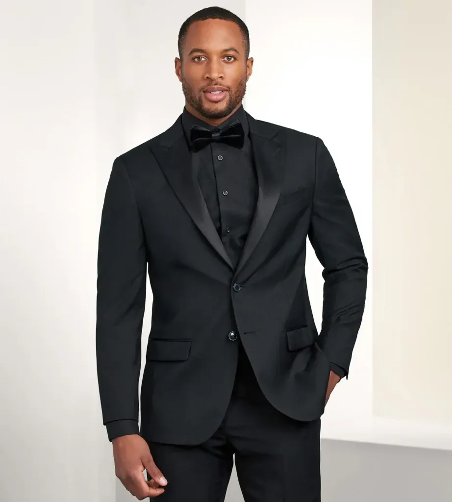 A groom wearing a sleek black tuxedo from Tip Top stands confidently in a well-lit room, showcasing attire perfect for a wedding. The tuxedo features a classic satin lapel and neatly tailored fit, complemented by a matching black bow tie and crisp black shirt, suitable for an elegant wedding setting. This image highlights Tip Top's offering in the wedding tuxedo category, emphasizing sophistication and style for grooms seeking a timeless look.