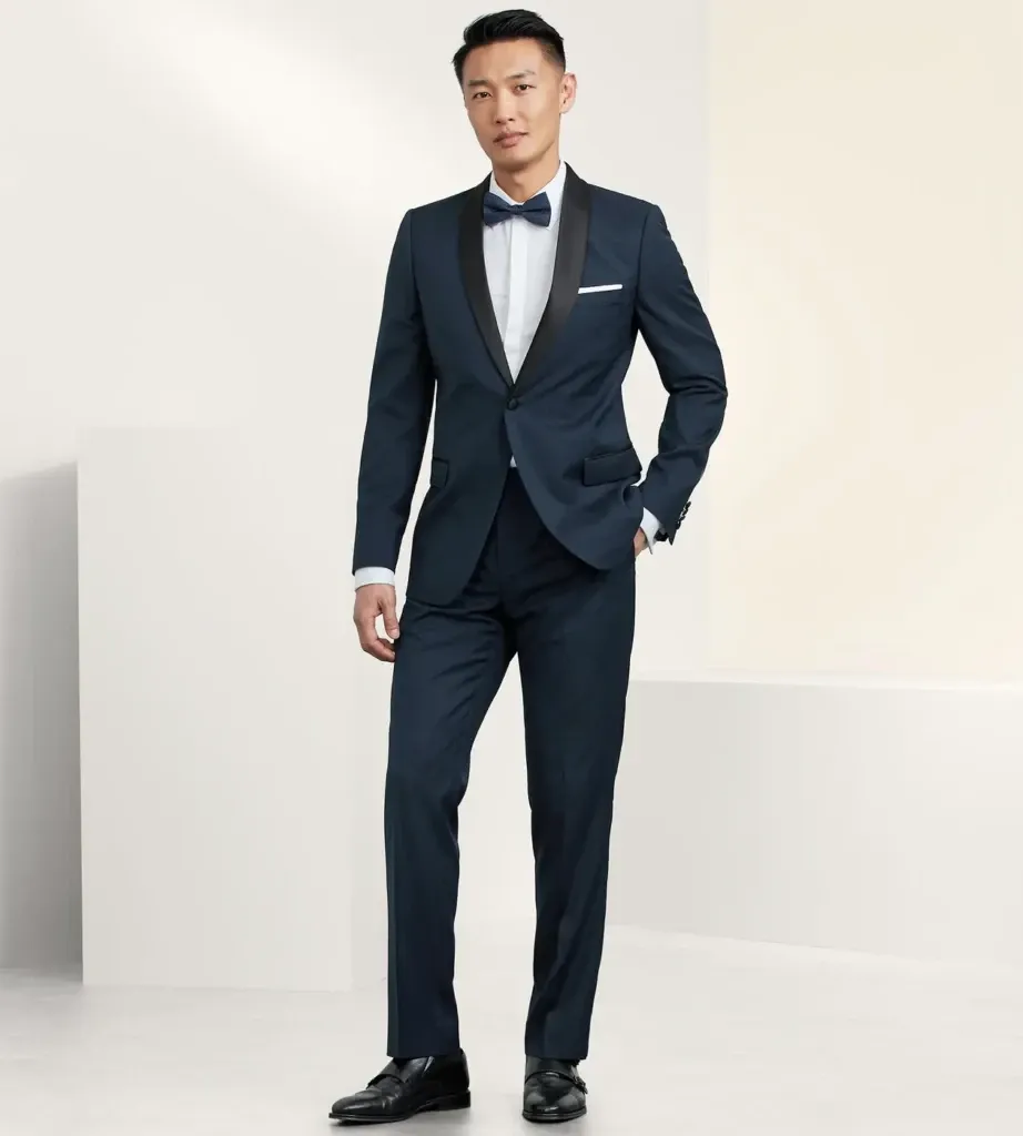 A groom is wearing an elegant navy tuxedo designed by Tip Top, categorized under Wedding Tuxedo. The tuxedo features a satin shawl lapel, paired with a crisp white dress shirt and a black bow tie. He also has a white pocket square and black dress shoes, completing the sophisticated wedding attire. The background is minimalistic, enhancing the focus on the stylish ensemble ideal for a formal wedding setting.