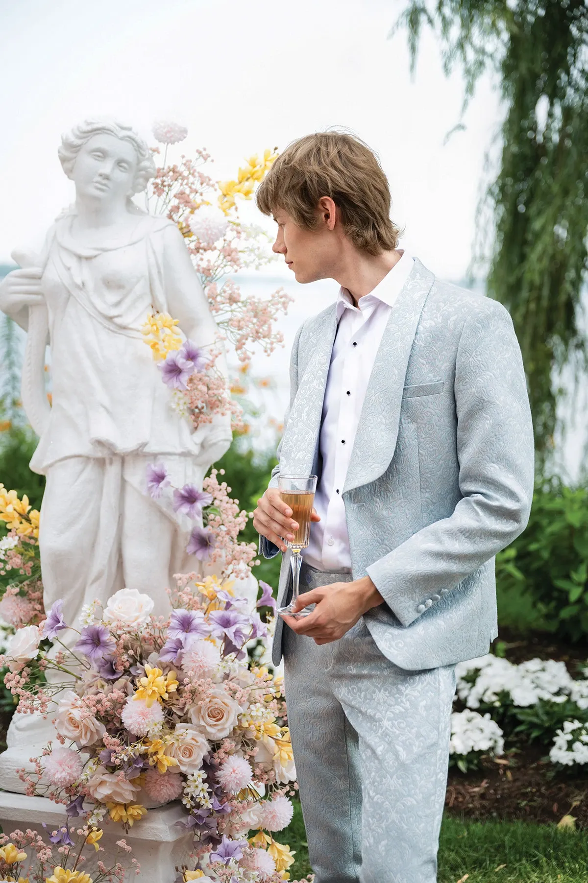 A groom stands elegantly beside a classical stone statue, surrounded by lush floral arrangements featuring pastel roses, lilacs, and yellow blooms, embodying the romantic essence of a wedding setting. He is dressed in a sophisticated light grey tuxedo with a subtle pattern from Jaco Uomo, highlighted by a crisp white dress shirt. Holding a champagne flute, he gazes thoughtfully, perfectly embodying the Wedding Tuxedo category. The image captures a refined, serene moment, ideal for showcasing Jaco Uomo's exquisite offerings on Mariage Elegant, Canada.