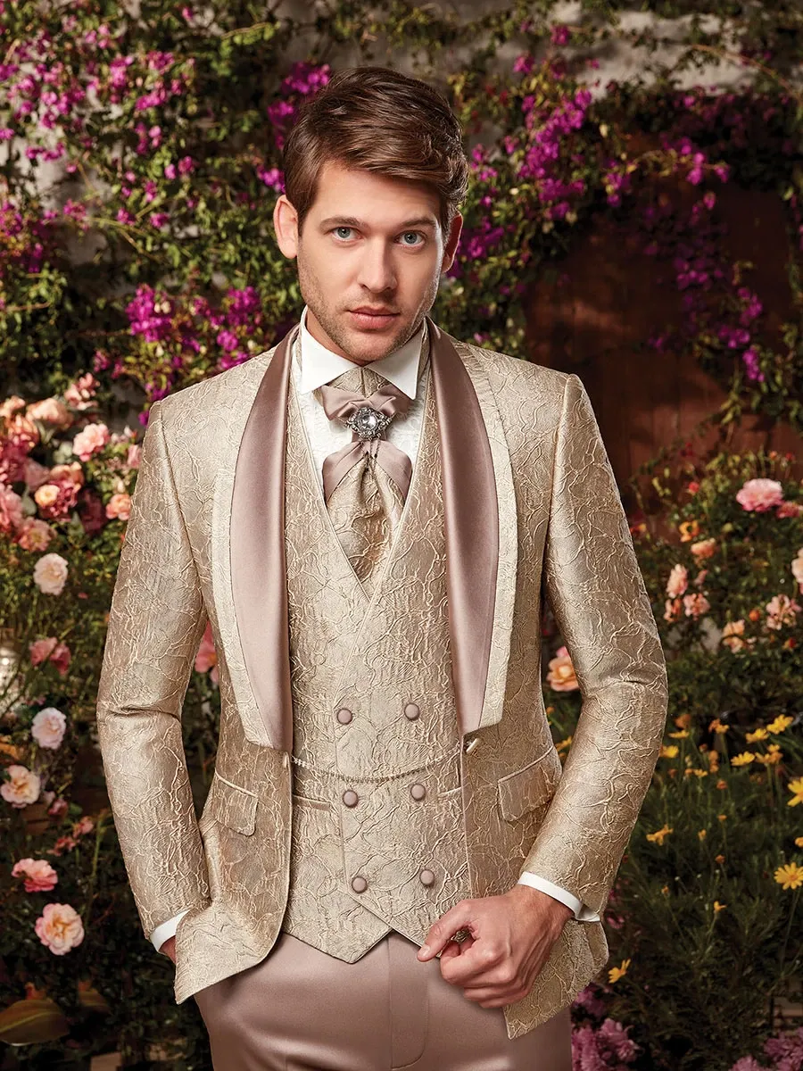 A man is wearing an elegant, golden-brown tuxedo by Jaco Uomo, perfectly tailored for a wedding. The tuxedo features a textured, embossed pattern with a satin shawl collar and matching waistcoat, adding a sophisticated touch. He pairs it with a white dress shirt and a distinctive, patterned cravat, ideal for a formal wedding setting. The backdrop is a lush garden adorned with vibrant, blooming flowers, providing a romantic and picturesque ambiance, suitable for a wedding. This image showcases Jaco Uomo as a premier choice in the wedding tuxedo category for Mariage Elegant in Canada.