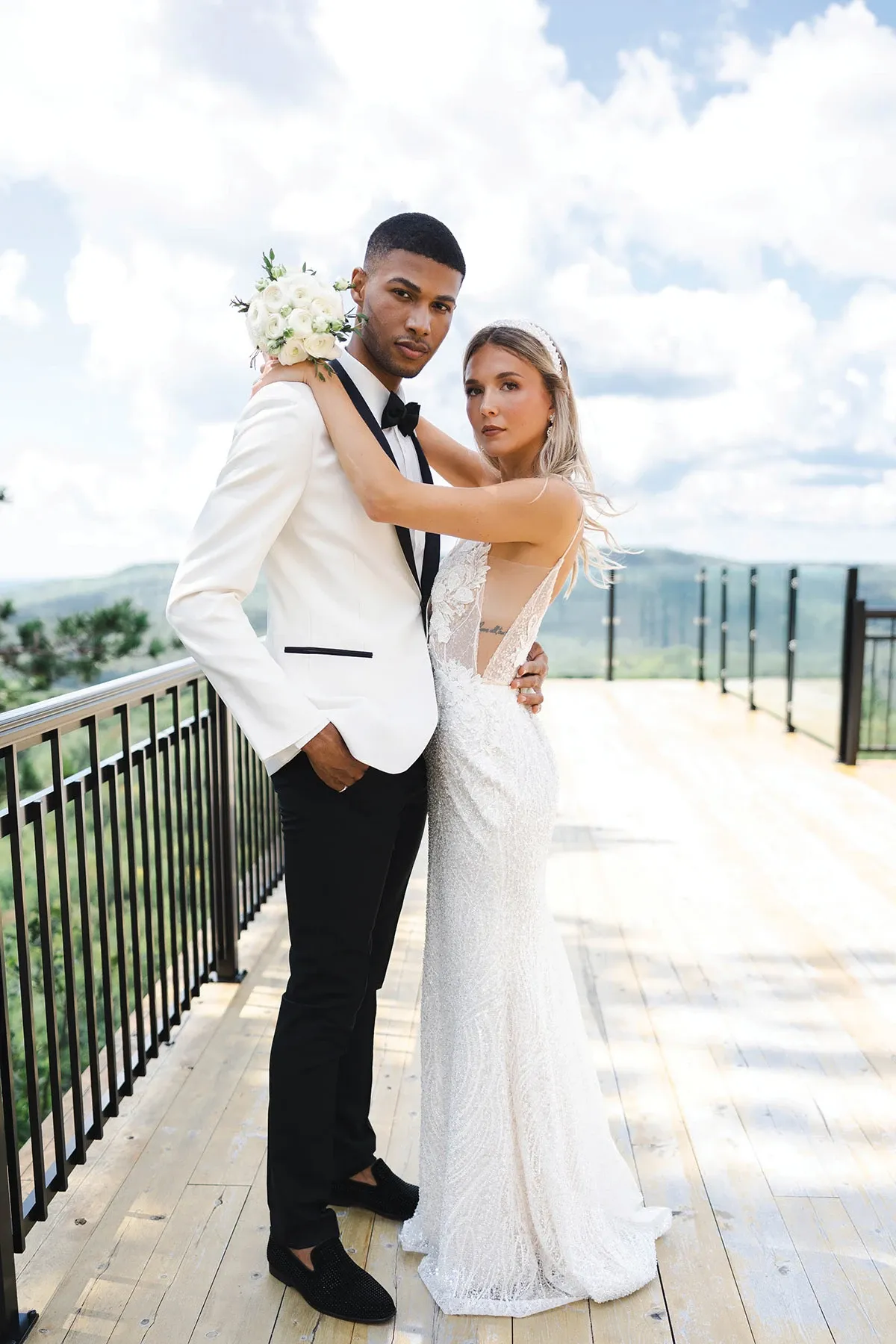 A couple stands elegantly on a scenic outdoor wooden platform with a mountainous backdrop. The groom is wearing a sophisticated tuxedo from Jaco Uomo, part of the wedding tuxedo collection. His attire features a crisp white jacket with black lapels, paired with black trousers and a classic black bow tie. The bride is wearing a stunning white lace wedding gown, holding a small bouquet of white flowers. The setting conveys an atmosphere of romance and elegance, perfect for a stylish wedding ceremony in Canada.