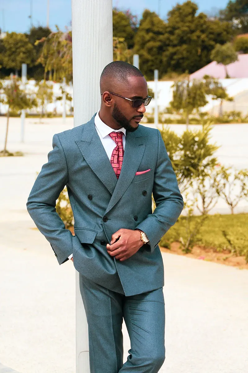A groom wearing a stylish, tailored grey tuxedo from Jaco Uomo, part of the Wedding Tuxedo category on Mariage Elegant, Canada. The tuxedo features a double-breasted design, paired with a crisp white shirt and a red patterned tie, adding a touch of elegance and sophistication. The groom accessorizes with sunglasses, a watch, and a pocket square, enhancing the modern look. The outdoor setting, with lush greenery and a bright, clear sky, suggests a chic wedding atmosphere.