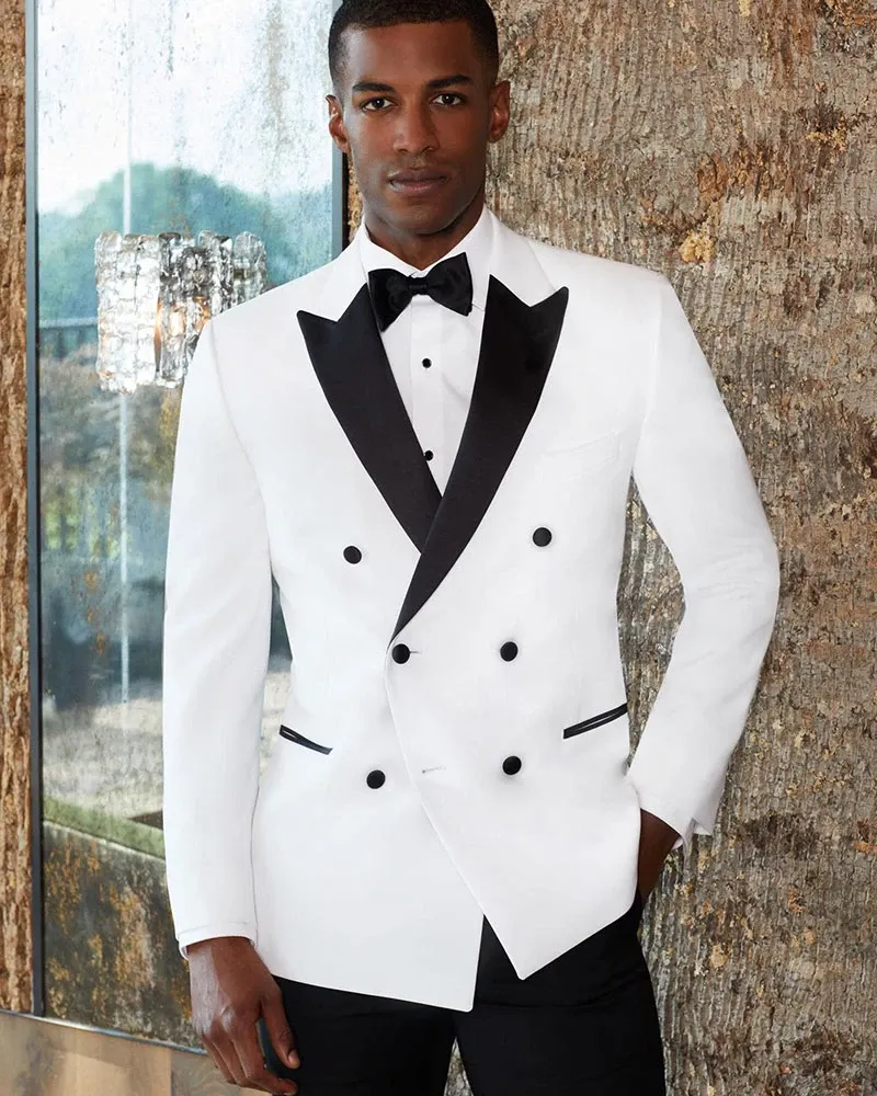 A groom is elegantly dressed in a sophisticated wedding tuxedo by Jaco Uomo, showcasing a sleek, double-breasted white jacket with black lapels. The tuxedo features black buttons and matching black pocket accents, paired with a classic black bowtie and crisp white dress shirt. The modern and stylish design is ideal for formal weddings, emphasizing elegance and refinement. The setting includes a textured, rustic wall and a glimpse of a large window, adding a blend of natural and contemporary elements to the scene. This image highlights Jaco Uomo as a premier vendor in the Wedding Tuxedo category for Mariage Elegant in Canada.