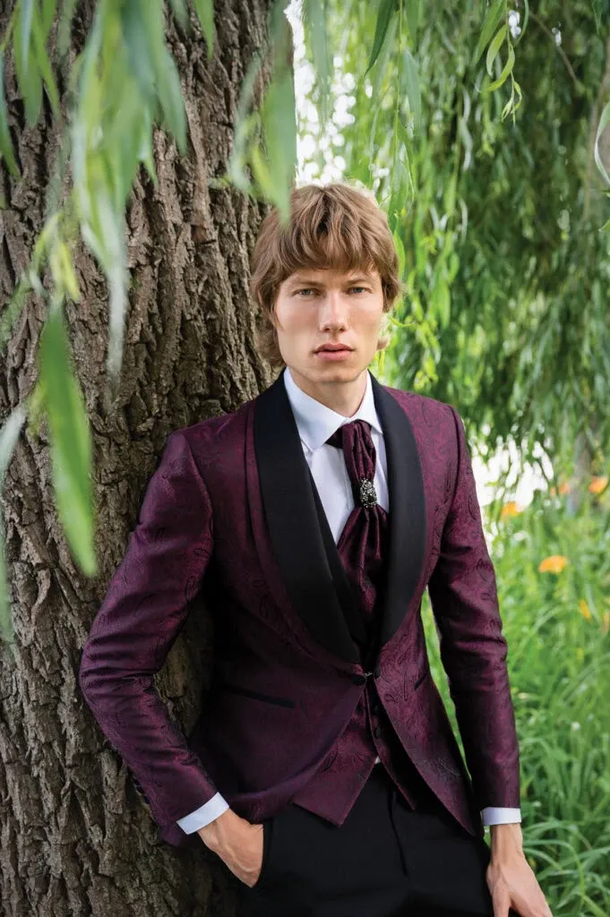 A groom is elegantly dressed in a sophisticated tuxedo by Jaco Uomo for a wedding occasion. He is wearing a rich burgundy tuxedo jacket with intricate floral patterns, paired with a black satin shawl lapel. The look is complemented by a white dress shirt and a burgundy cravat, fastened by a silver brooch. He is standing against a tree, surrounded by lush greenery, providing a serene and natural backdrop that enhances the formal attire. This image highlights Jaco Uomo's expertise in wedding tuxedos, perfect for an elegant and stylish wedding in Canada, featured on Mariage Elegant.