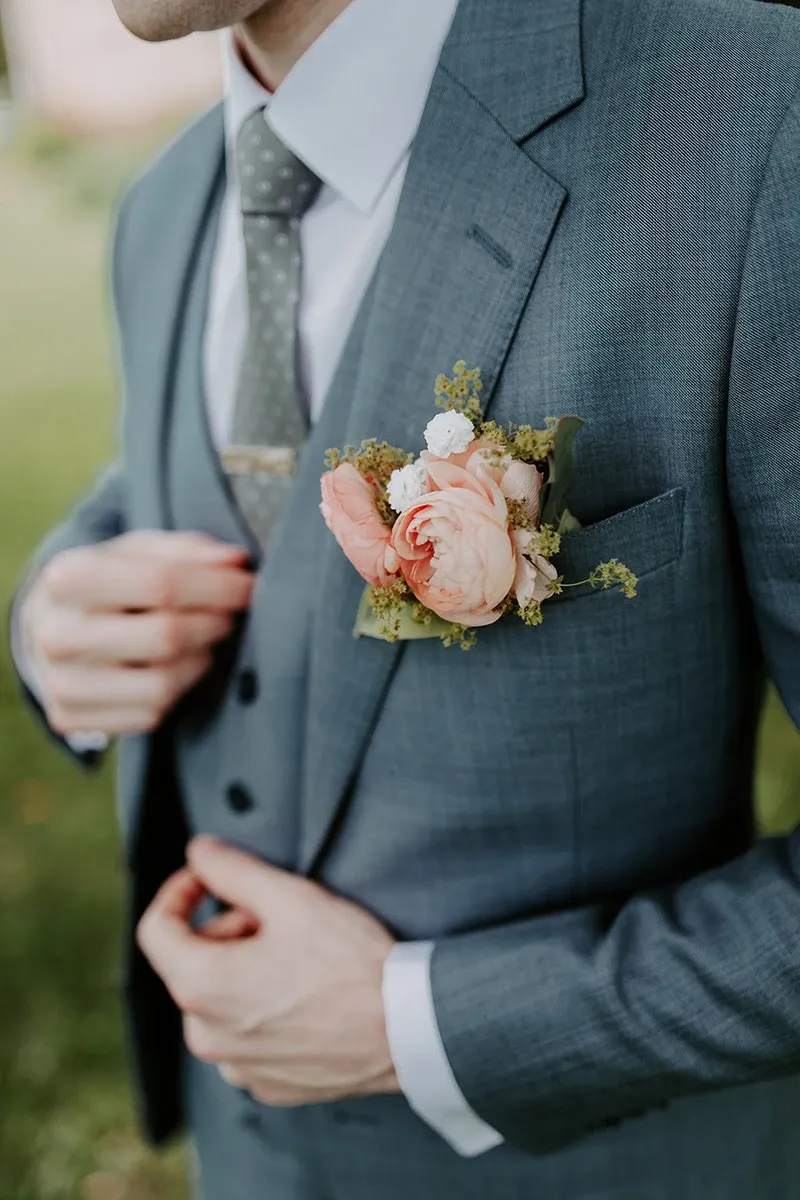 A groom is elegantly dressed in a tailored, light gray three-piece suit from Jaco Uomo, under the wedding tuxedo category. The suit features a well-fitted jacket, with a subtle check pattern, complemented by a white dress shirt and a dotted gray tie. The gentleman is adjusting his tie, creating a sense of anticipation and readiness for the wedding ceremony. In the breast pocket of the suit, there is a beautifully arranged boutonniere with soft pink roses and delicate greenery, adding a touch of elegance and romance to the attire. The image emphasizes the quality and style offered by Jaco Uomo, ideal for grooms seeking sophisticated wedding apparel.