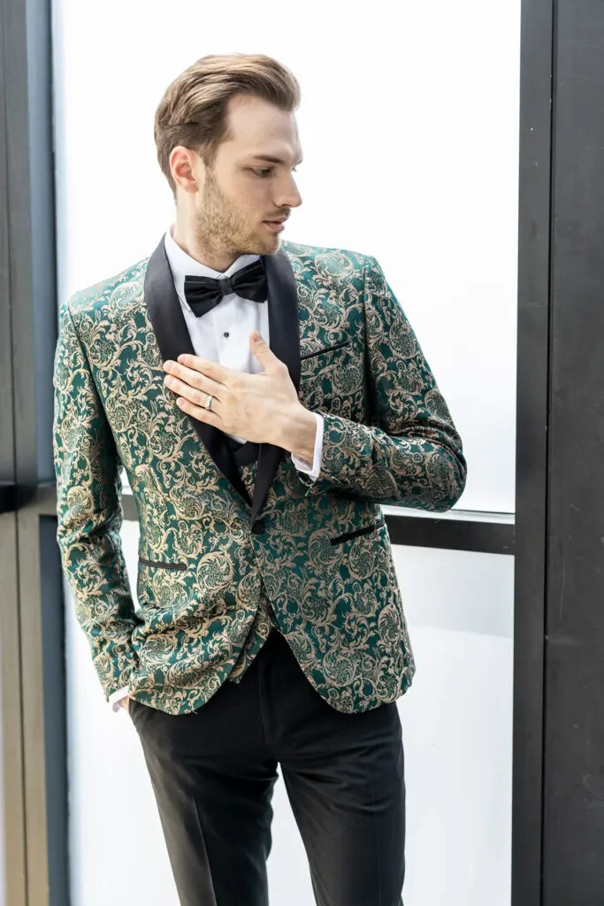 A groom elegantly poses indoors wearing a distinguished green and gold paisley tuxedo designed by Jaco Uomo, a vendor in the Wedding Tuxedo category for Mariage Elegant, Canada. The tuxedo features a sleek black satin lapel and is complemented by a classic white dress shirt and a black bow tie. The outfit is paired with tailored black trousers, showcasing a sophisticated style perfect for a wedding setting. The groom's confident stance and the tuxedo's intricate detailing contribute to a luxurious and refined bridal look.