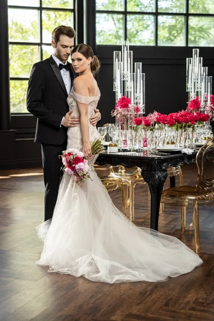In this elegant wedding scene from Mariage Elegant, Canada, the couple stands gracefully near a beautifully set dining table adorned with tall, crystal candle holders and vibrant pink floral arrangements. The groom is wearing a classic black tuxedo from Jaco Uomo, fitting the wedding tuxedo category perfectly with its sophisticated style. The bride, dressed in a flowing white gown, holds a bouquet of matching pink flowers, complementing the romantic and refined atmosphere. Large windows in the background allow natural light to illuminate the room, enhancing the overall luxurious ambiance of the setting.