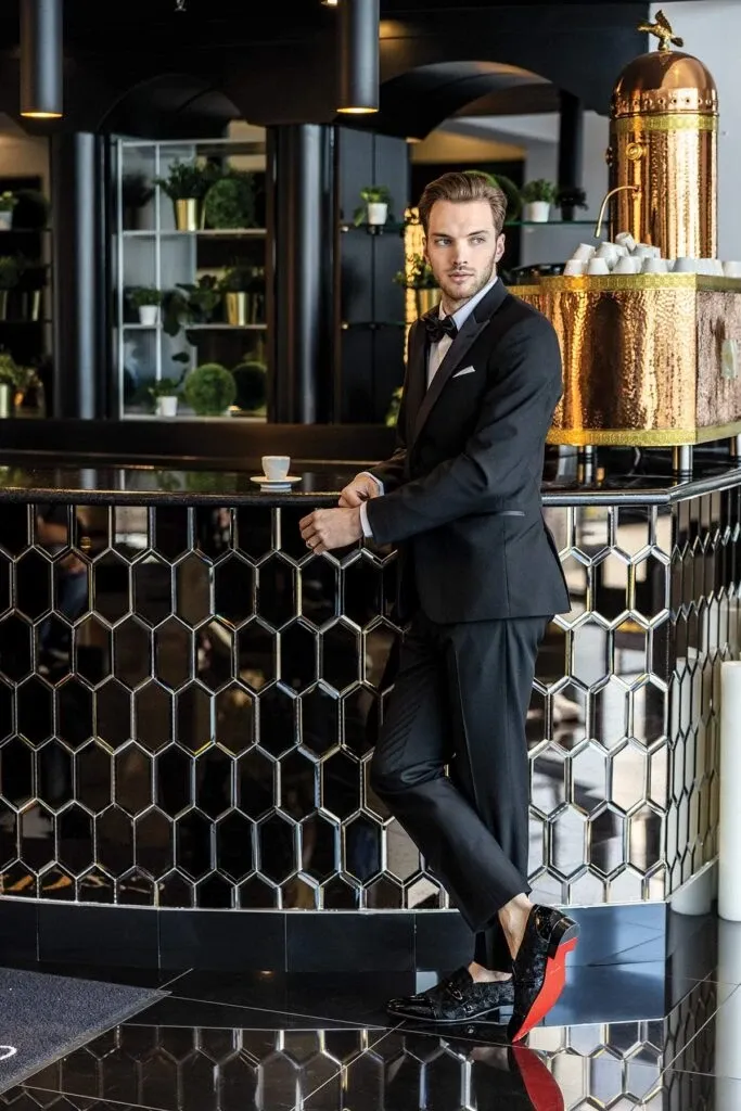 A groom is elegantly posed in a sophisticated setting, wearing a stylish tuxedo from Jaco Uomo, part of the Wedding Tuxedo collection. The tuxedo features a classic black design with satin lapels and is paired with a crisp white shirt and a black bow tie, creating a timeless wedding look. The setting includes a modern, polished counter with honeycomb tiles and lush greenery in the background, contributing to a luxurious and celebratory atmosphere. This image is ideal for showcasing formal wedding attire options available through the vendor Jaco Uomo at Mariage Elegant in Canada.