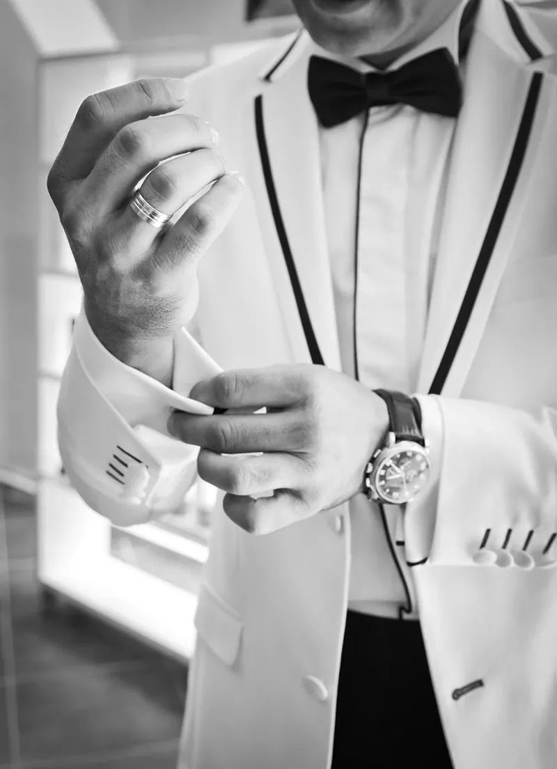 A groom adjusts his stylish cufflinks on a crisp, white tuxedo by Jaco Uomo, featured in the wedding tuxedo category. The tuxedo is impeccably tailored with sleek black trim, complemented by a classic black bow tie. A sophisticated wristwatch and a dual-band ring add elegance to the ensemble, ideal for a sophisticated wedding celebration. The setting, captured in black and white, highlights the timeless and elegant atmosphere associated with Mariage Elegant in Canada.