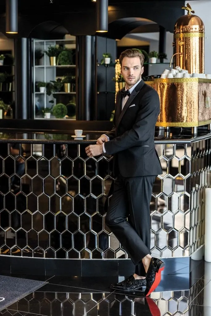A man stands confidently by a modern bar counter, adorned with a sophisticated honeycomb pattern, wearing an elegant black tuxedo from Jaco Uomo under the wedding tuxedo category. The tuxedo features a classic bow tie and a crisp white shirt, highlighting the vendor’s commitment to high-quality wedding attire. The setting includes luxurious decor with a sleek, reflective floor and artistic design elements, complementing the formal ambiance suitable for wedding events.
