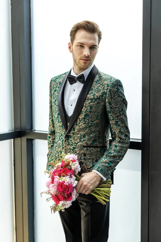 A groom is standing by a large window dressed in an elegant tuxedo by Jaco Uomo, a featured wedding tuxedo vendor on Mariage Elegant, Canada. He is wearing a striking green and gold brocade jacket with a classic black satin lapel over a crisp white dress shirt complemented by a black bow tie. The ensemble is completed with black trousers. In his hand, he holds a vibrant bouquet of pink and red flowers, adding a touch of romance and color to the sophisticated attire, perfectly suited for a stylish and modern wedding.