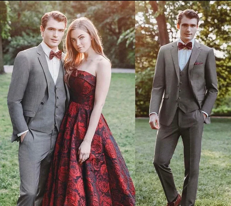A couple poses elegantly outdoors, showcasing formal wedding attire. The groom is wearing a stylish gray tuxedo from Jaco Uomo, a vendor in the Wedding Tuxedo category, complemented by a white dress shirt and a maroon bow tie, perfect for a classic wedding setting. Beside him, the bride stands gracefully in a strapless gown with a black and red floral pattern. In another image, the groom stands alone, emphasizing the tailored fit of his three-piece suit, including a vest, enhancing the sophisticated wedding theme offered by Jaco Uomo, part of Mariage Elegant in Canada.