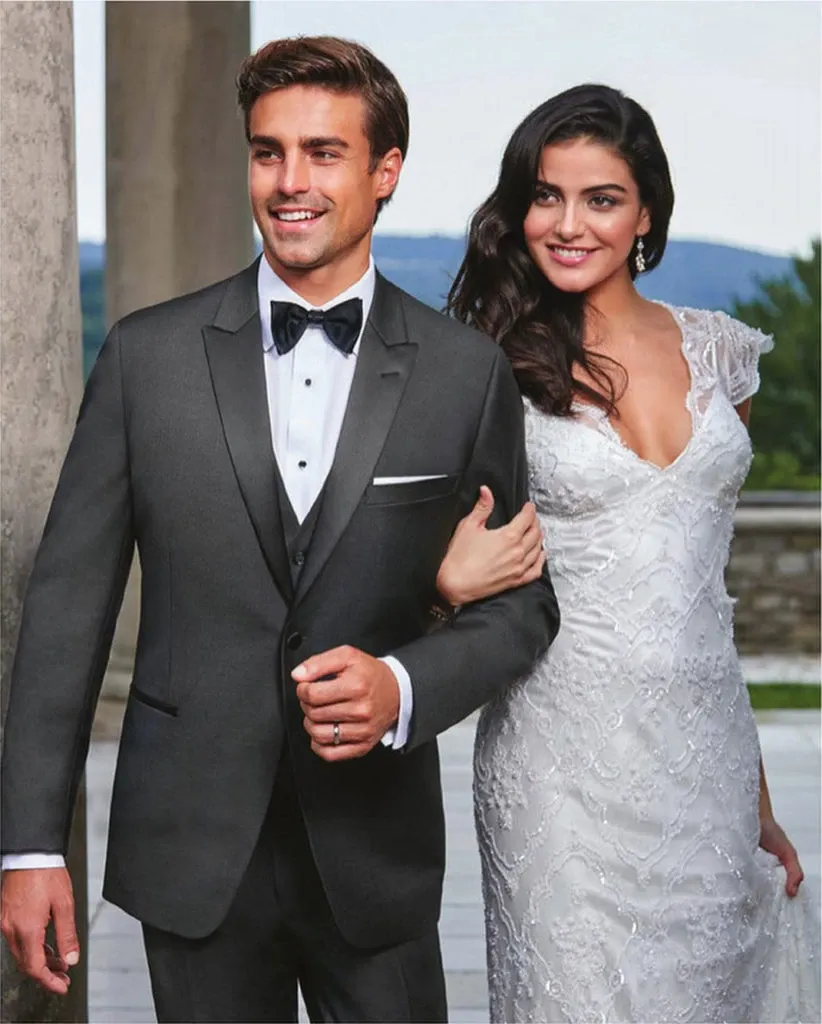 A groom wearing an elegant black tuxedo from Jaco Uomo is standing next to a bride in a stunning white lace wedding gown. The groom's tuxedo features a classic bow tie and a crisp white dress shirt, epitomizing sophistication and style within the wedding tuxedo category. They are linked arm-in-arm, smiling, and looking towards the distance, set against a backdrop of stone pillars and a scenic view, suggesting a serene wedding venue. This image reflects the high-quality offerings of Mariage Elegant's vendor listings in Canada, showcasing formal attire perfect for a memorable wedding day.