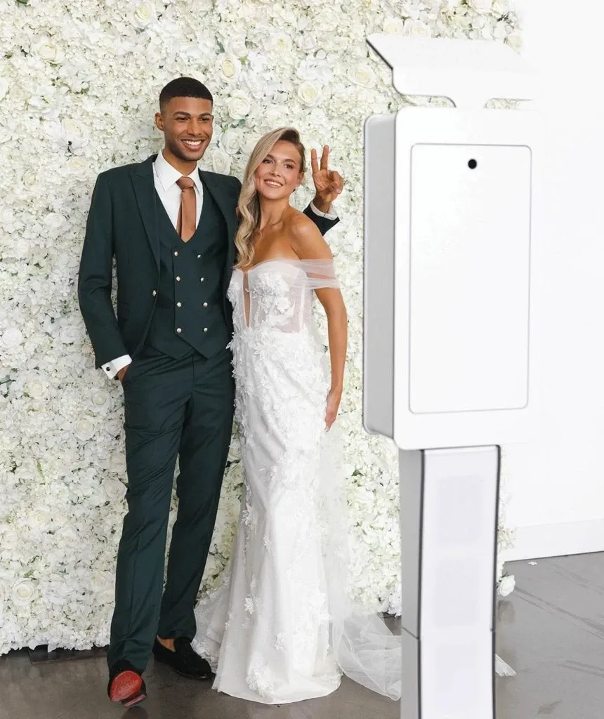 A newlywed couple poses joyfully in front of a stunning white floral backdrop. The groom is dressed in a sophisticated dark green tuxedo by Jaco Uomo, perfectly fitting the elegant wedding category. He complements his look with a stylish brown tie and polished brown shoes. The bride stands beside him, radiant in an exquisite white lace gown with off-the-shoulder sleeves that adds a touch of elegance to the scene. She playfully flashes a peace sign. In the foreground, a sleek photo booth is visible, hinting at a modern and interactive wedding reception. The setting exudes charm and contemporary wedding style, ideal for those seeking inspiration through Mariage Elegant in Canada.