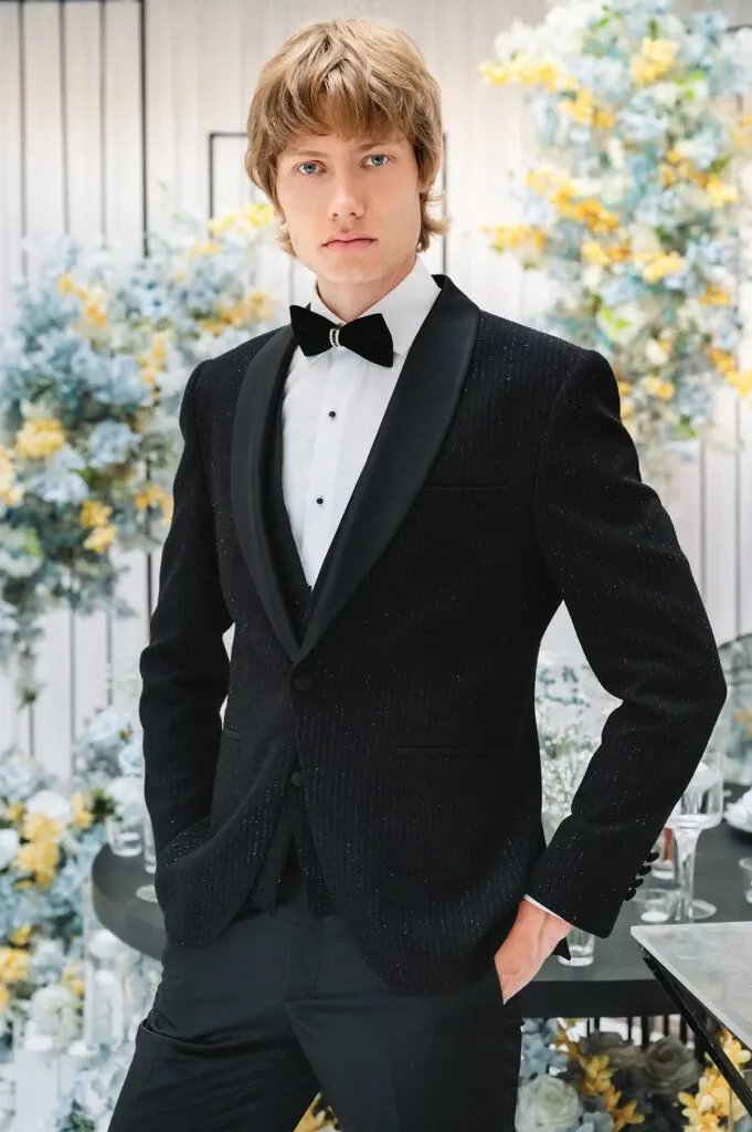 This image features a model wearing a classic black tuxedo from vendor Jaco Uomo, part of the Wedding Tuxedo category on Mariage Elegant, Canada. The tuxedo is tailored with a sleek black jacket adorned with subtle pinstripes, paired with a crisp white dress shirt and a black bow tie. The setting is elegant, with floral arrangements in soft blues and yellows visible in the background, creating a sophisticated wedding atmosphere. The overall look exemplifies refined style, perfect for a formal wedding event.