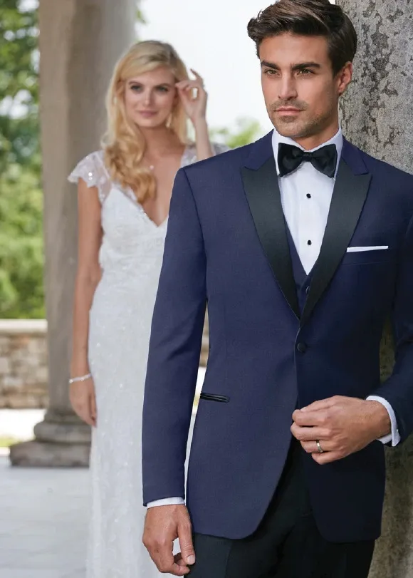 A groom stands confidently in an elegant navy tuxedo designed by Jaco Uomo, a vendor specializing in wedding tuxedos, featured on Mariage Elegant in Canada. The tuxedo is paired with a classic black bow tie and a crisp white shirt, sharply contrasting with the lush green backdrop. In the background, a bride in a delicately detailed white gown with lace cap sleeves gazes at him lovingly, exemplifying a perfect wedding day moment. The image captures the sophisticated attire options available for grooms through Jaco Uomo, highlighting the elegance and style suitable for a wedding setting.