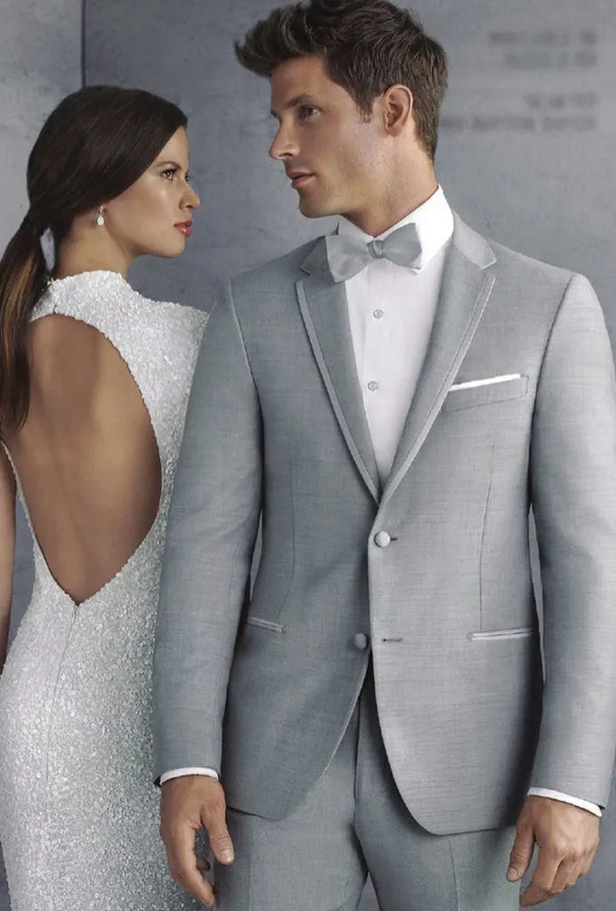 A groom is elegantly dressed in a light grey tuxedo by Jaco Uomo, perfectly tailored for a wedding ceremony. The tuxedo features a classic fit with a single-breasted jacket and white dress shirt, complemented by a stylish bow tie and matching pocket square. Standing beside him is a bride wearing a shimmering white gown with a dramatic open-back design, adding a graceful and sophisticated touch. This image showcases the refined options offered by Jaco Uomo in the Wedding Tuxedo category, ideal for couples seeking stylish attire for their special day at Mariage Elegant in Canada.