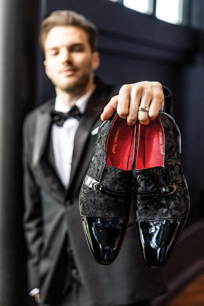 A groom holding a pair of elegant black tuxedo shoes with a glossy finish and intricate lace details, designed by Jaco Uomo. He wears a sleek black tuxedo with a classic white dress shirt and a black bow tie, fitting the category of Wedding Tuxedo. The setting exudes a sophisticated, upscale ambiance, appropriate for a modern wedding. The groom's wedding band is visible, adding a personal touch and highlighting the significance of the occasion. This image showcases the high-quality, stylish offerings of Jaco Uomo for weddings listed on Mariage Elegant, Canada.