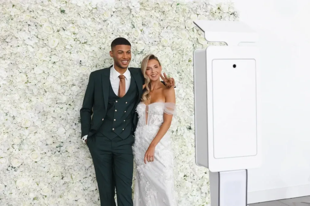 This image features a couple posing in front of a wall adorned with white flowers. The groom is wearing a stylish dark green tuxedo by Jaco Uomo, fitting the wedding tuxedo category, which adds a modern and elegant flair to the occasion. The bride is in a stunning, off-the-shoulder white gown, complementing the groom's attire. A sleek photo booth is visible in the foreground, suggesting a fun, interactive element for wedding guests. The overall setting exudes an air of sophistication and romance, ideal for a memorable wedding celebration in Canada.