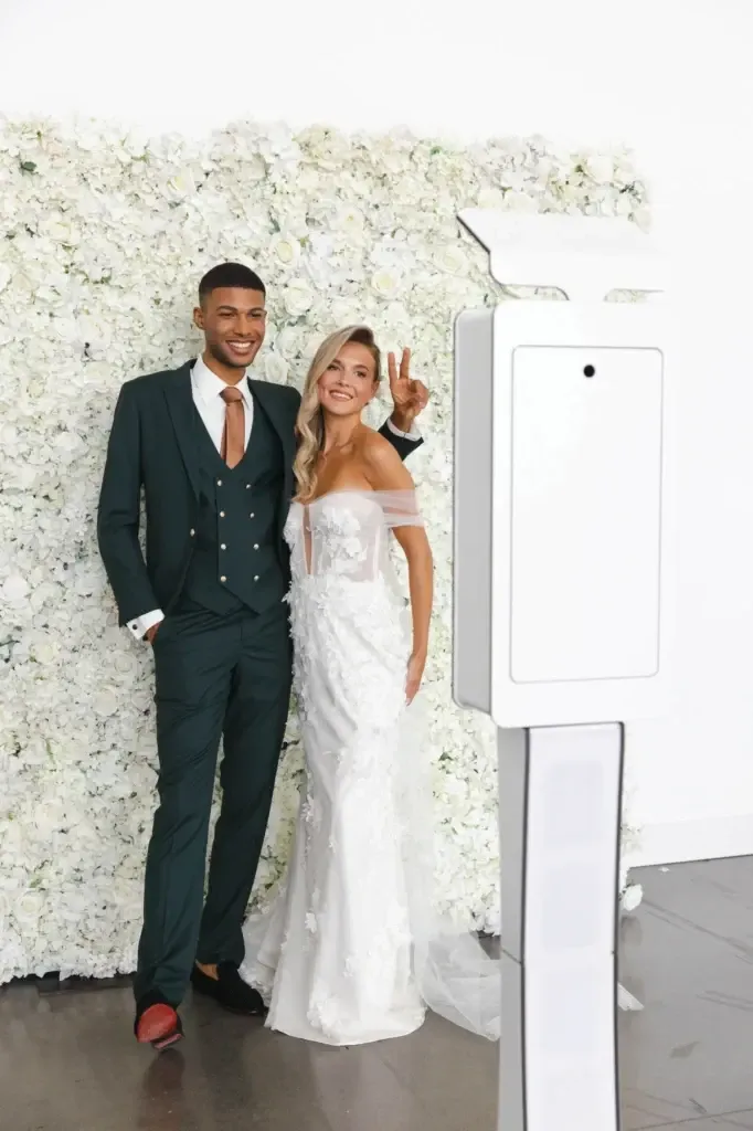 A smiling couple stands in front of a lush floral backdrop covered in white flowers, showcasing elegant wedding attire. The groom is wearing a sophisticated, dark green tuxedo from Jaco Uomo, paired with a crisp white shirt and black dress shoes, fitting the Wedding Tuxedo category. The bride is in a stunning off-the-shoulder white gown with delicate lace detailing. In the foreground, a modern photo booth is visible, capturing the joyful moment. This setup is perfect for weddings and showcases the refined offerings of Mariage Elegant in Canada.