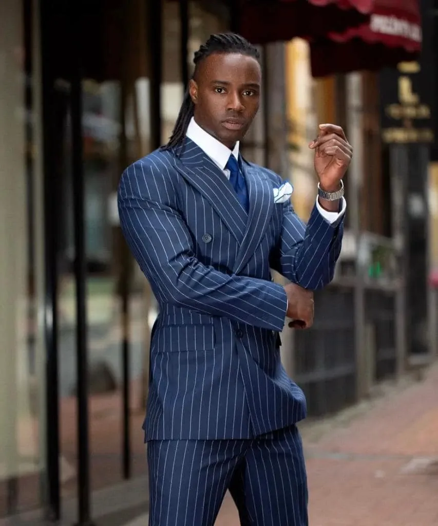 A man stands confidently on a city street, wearing a sophisticated, double-breasted, blue pinstripe tuxedo designed by Jaco Uomo. The tuxedo is complemented by a crisp white pocket square and a navy blue tie, suitable for a wedding environment. He is posing with one hand adjusting his sleeve, showcasing the elegant tailoring of the outfit. The backdrop features an urban setting with blurred storefronts, creating a contrast that highlights the formal attire. This image represents the Wedding Tuxedo category for Mariage Elegant in Canada, ideal for grooms seeking a modern and stylish look.