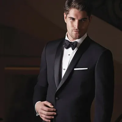 A groom is elegantly dressed in a classic black tuxedo provided by Jaco Uomo, a prestigious wedding tuxedo vendor. The tuxedo features a sleek satin lapel and a crisp white shirt underneath. The ensemble is complemented by a black bow tie and a white pocket square, adding a touch of sophistication. This attire, showcased in a refined indoor setting, is ideal for a timeless and elegant wedding celebration, reflecting the high standards of Mariage Elegant in Canada.