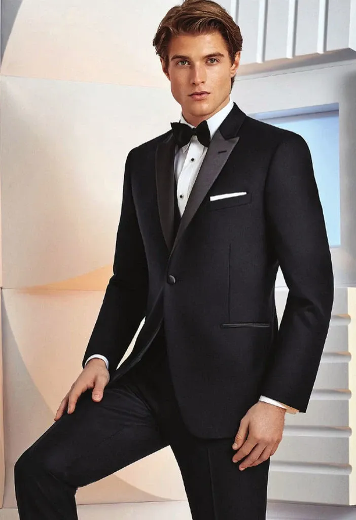 A groom poses elegantly in a sleek black tuxedo by Jaco Uomo, perfect for a wedding setting. The tuxedo features a satin lapel and a single-breasted design, complemented by a crisp white dress shirt and a classic black bow tie. The model stands against a modern, geometric backdrop, embodying sophistication, making this a distinguished choice for wedding attire. This image is featured in the Wedding Tuxedo section of Mariage Elegant's vendor listings in Canada.