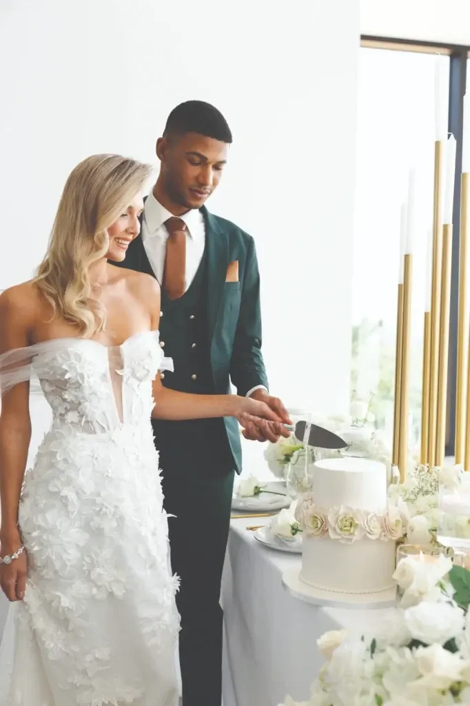 A couple elegantly stands together, both smiling as they cut a sophisticated white wedding cake. The groom is wearing a stylish, dark green tuxedo with a black lapel, designed by Jaco Uomo, a vendor specializing in wedding tuxedos. His ensemble is complemented by a crisp white shirt and a coordinating vest and tie. The bride is dressed in a stunning off-the-shoulder white wedding gown adorned with floral appliques. They are surrounded by a beautifully decorated table displaying white roses and candles, creating an elegant and romantic ambiance typical of a well-planned wedding event by Mariage Elegant in Canada.