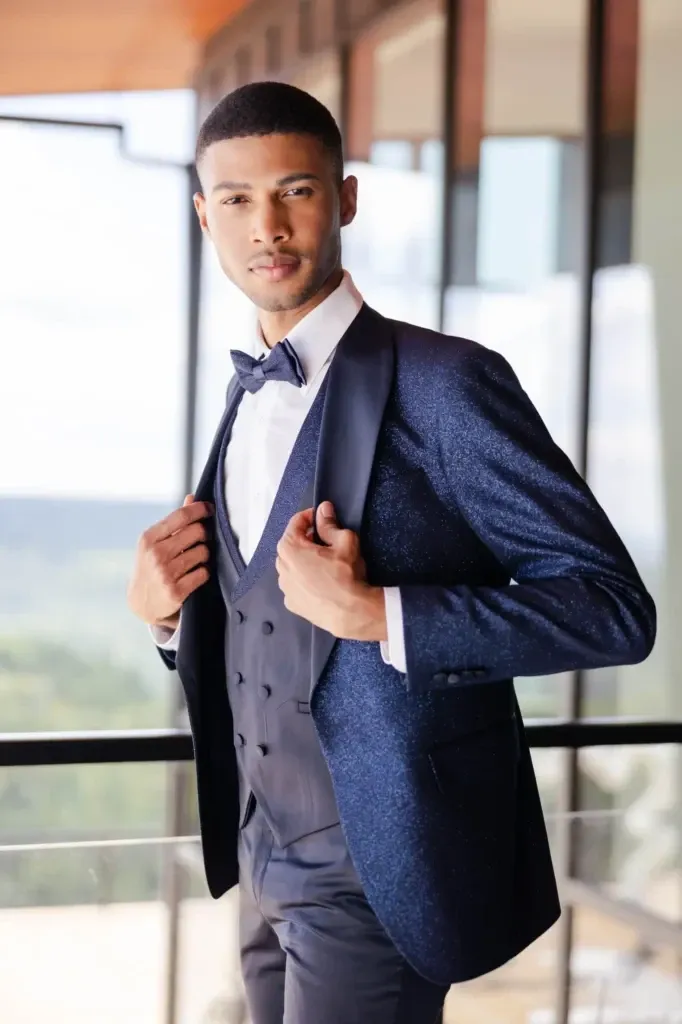 A groom is elegantly dressed in a tailored dark blue tuxedo with a subtle shimmer, provided by Jaco Uomo under the wedding tuxedo category for Mariage Elegant in Canada. The ensemble includes a matching waistcoat, a crisp white shirt, and a classic bow tie. The groom is adjusting his jacket while standing on a modern balcony with large glass windows, offering a view of lush greenery in the distance, creating a sophisticated and stylish wedding look.